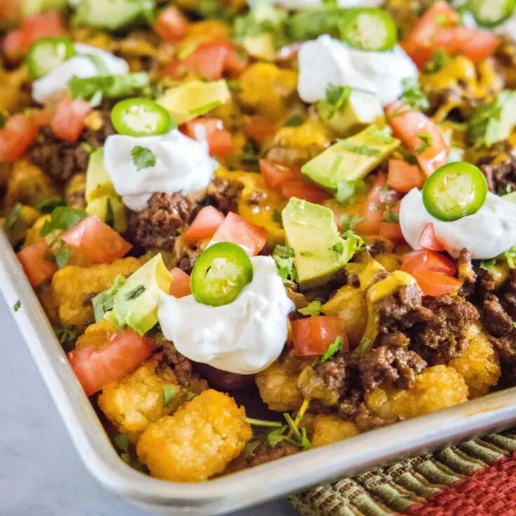 The corner of a baking sheet with ground beef tater tot nachos, topped with jalapenos, sour cream, tomatoes, avocado, and cilantro