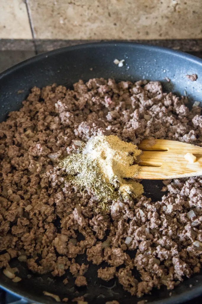 Ground beef in a skillet with garlic and onion powder on top, with a wooden spoon.