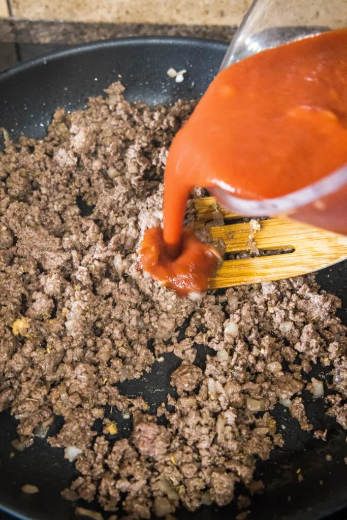 Tomato sauce being poured into a skillet of ground beef, with a wooden spoon.