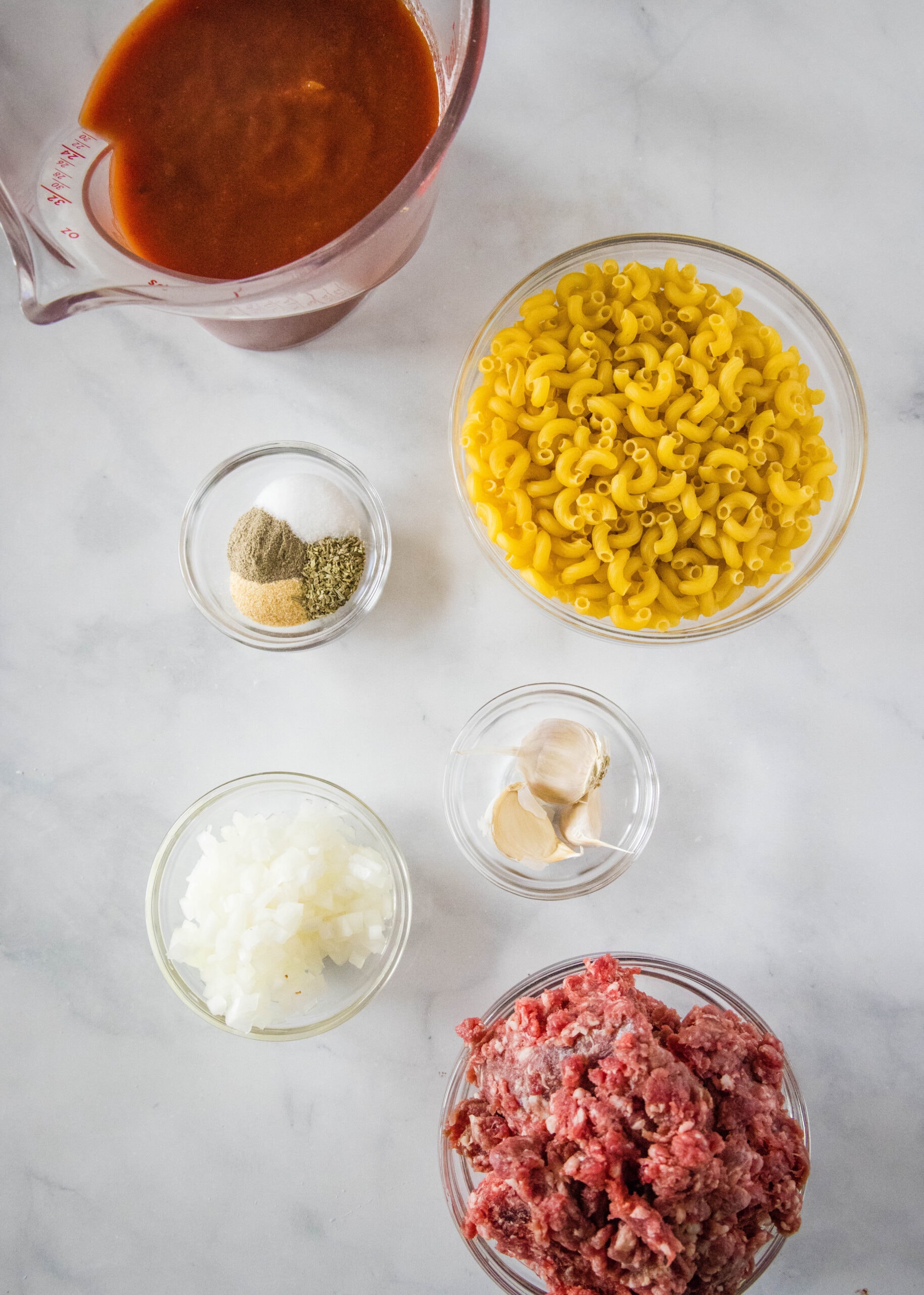 Overhead view of the ingredients needed for beefaroni: a bowl of macaroni, a bowl of ground beef, a pitcher of tomato sauce, a bowl of seasonings, a bowl of garlic, and a bowl of onions