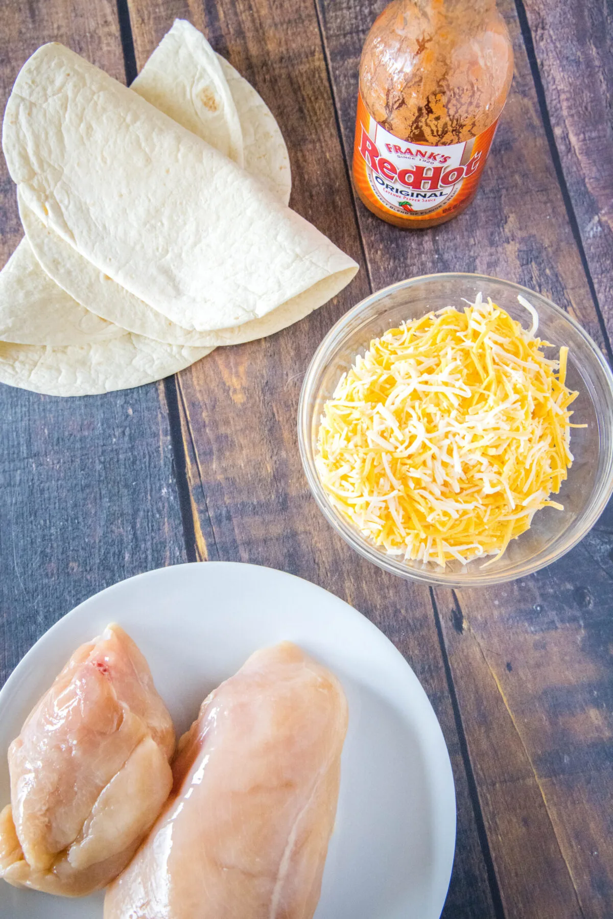 Overhead view of a plate of chicken breasts, a bowl of shredded cheese, a bottle of wing sauce, and two flour tortillas