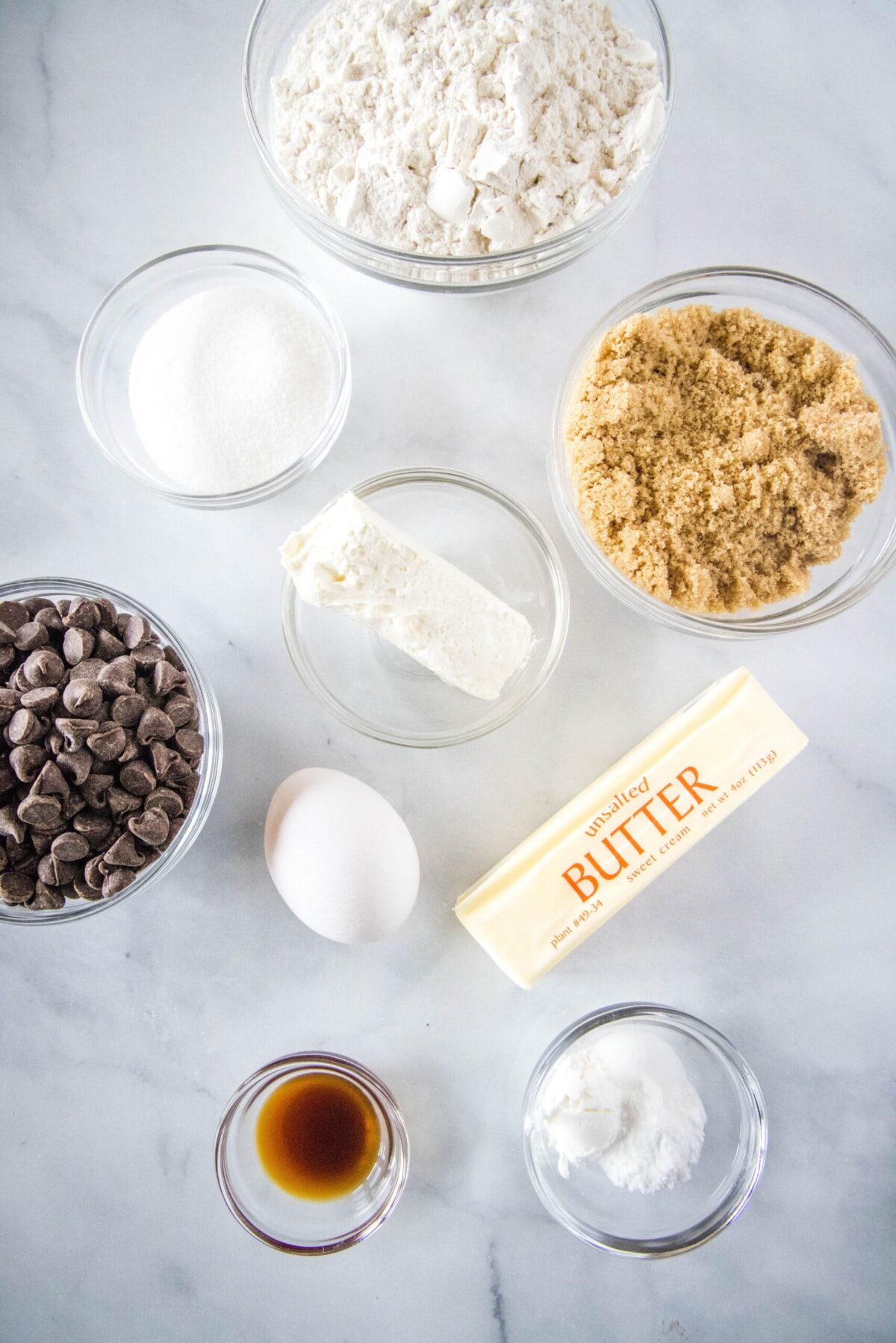 Overhead view of the ingredients needed for cream cheese chocolate chip cookies: a bowl of flour, a bowl of sugar, a bowl of chocolate chips, a bowl of brown sugar, a bowl of cream cheese, a bowl of vanilla extract, a bowl of baking soda and cornstarch, a stick of butter, and an egg