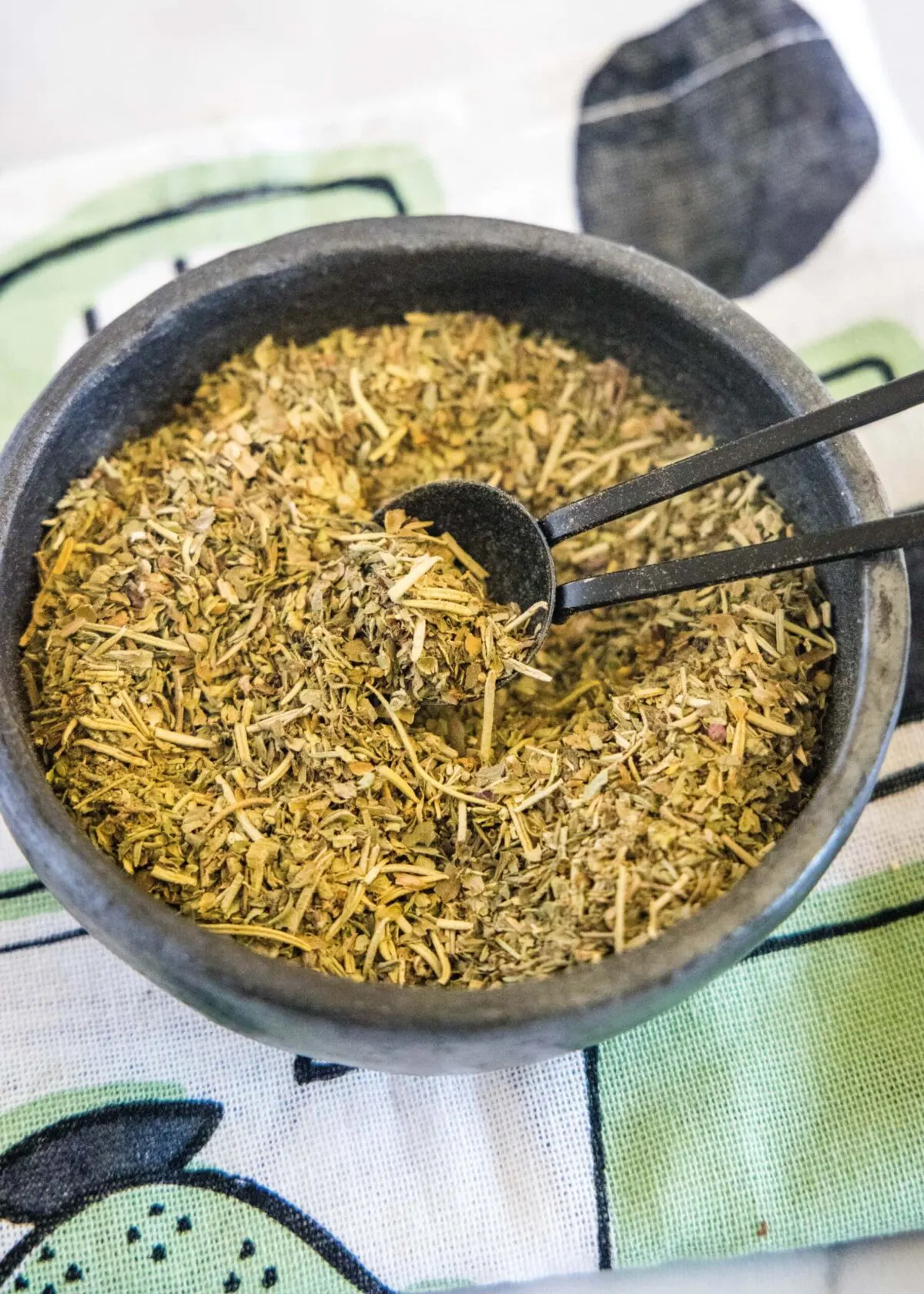 Close up overhead view of a bowl of Italian seasoning with a spoon in it, on a kitchen towel