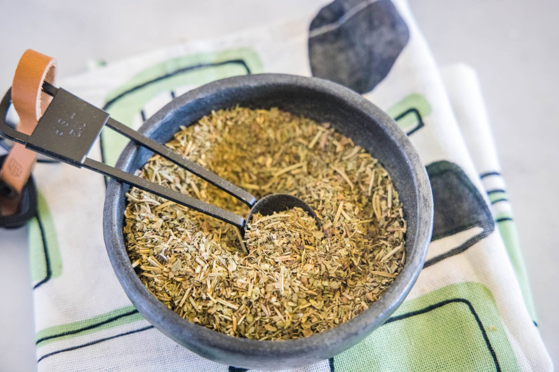Overhead view of a bowl of Italian seasoning with a spoon in it, on top of a kitchen towel