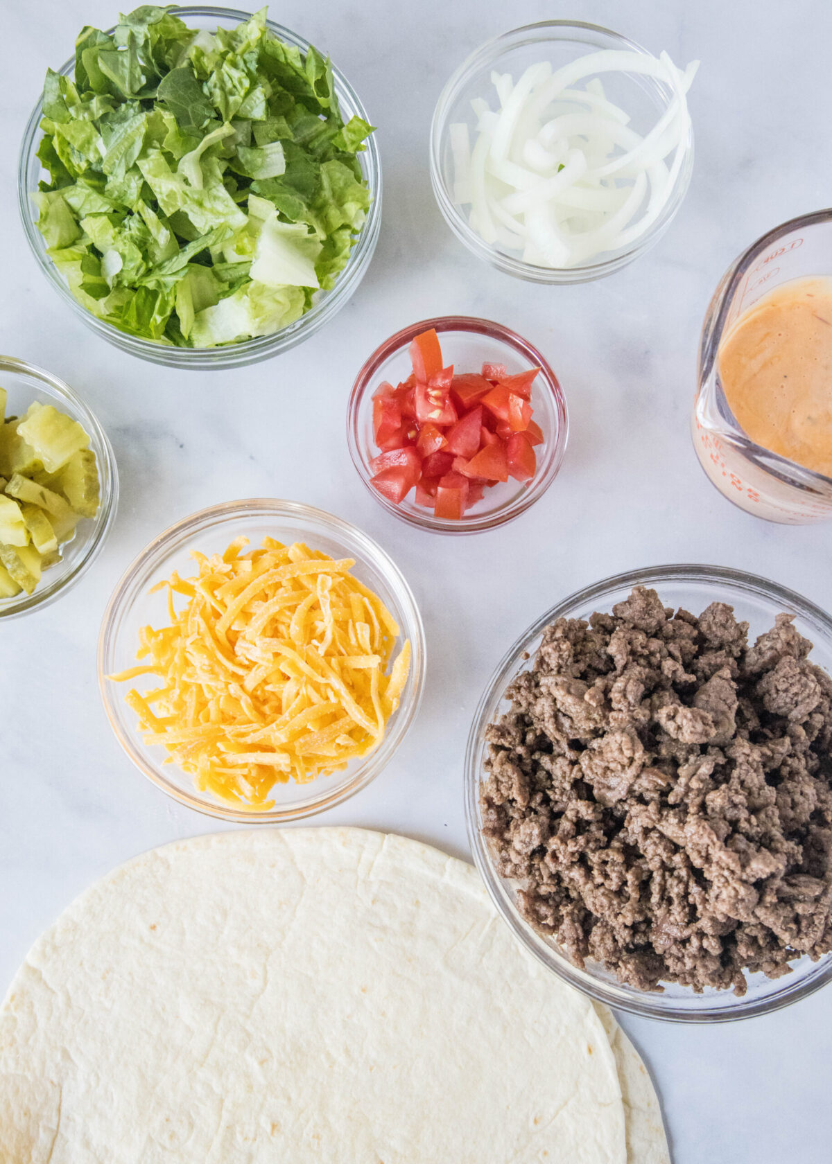 Overhead view of the ingredients needed for Big Mac wraps: a stack of tortillas, a bowl of cooked ground beef, a bowl of shredded cheese, a bowl of onions, a bowl of tomatoes, a bowl of pickles, a bowl of lettuce, and a jar of sauce