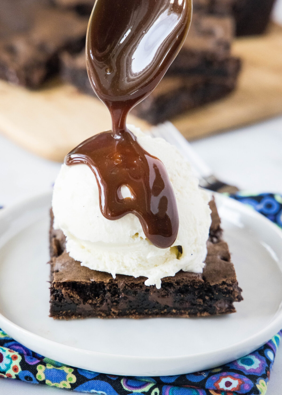 A spoon drizzling fudge sauce on top of ice cream on a square of brownie on a plate