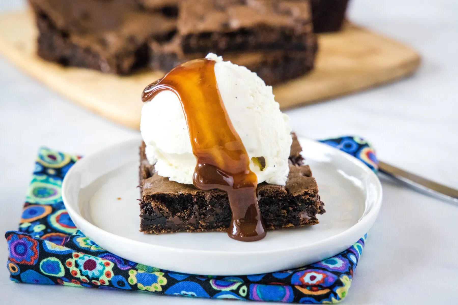 A plate with a brownie square, with a scoop of vanilla ice cream on top, with chocolate sauce dripping off it, with brownies in the background