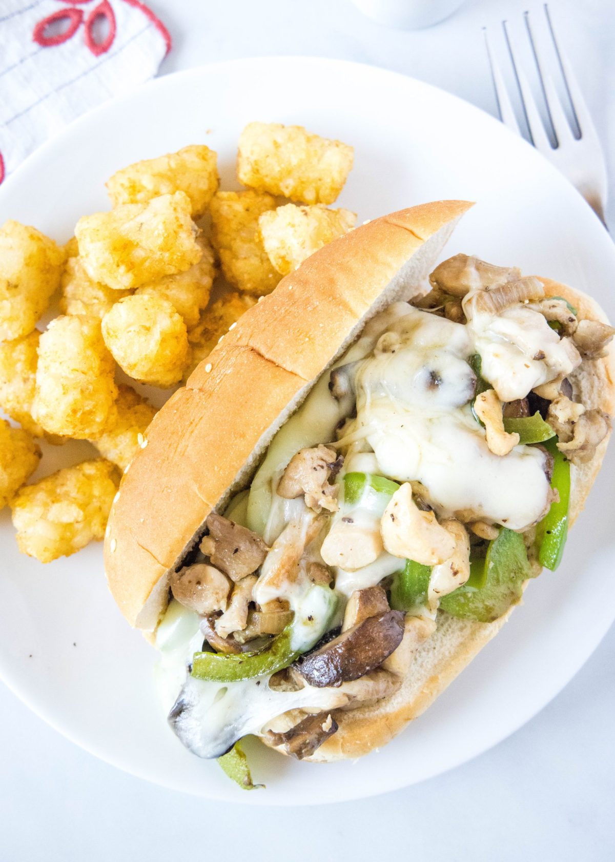 Overhead view of a chicken Philly cheesesteak on a plate next to some tater tots, with a fork on the side