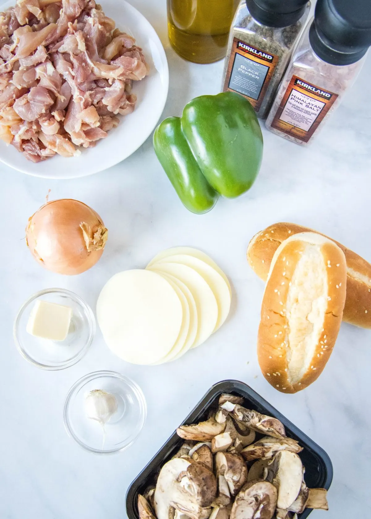 Overhead view of the ingredients you need for chicken Philly cheesesteaks: a bowl of sliced chicken, a green bell pepper, two hoagie rolls, a tub of mushrooms, an onion, slices of provolone cheese, a bowl of garlic, a bowl of butter, a jar of olive oil, a container of salt, and a container of pepper