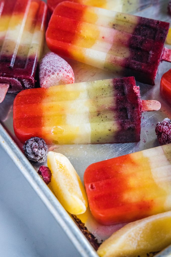 rainbow popsicles on a baking tray next to frozen fruit