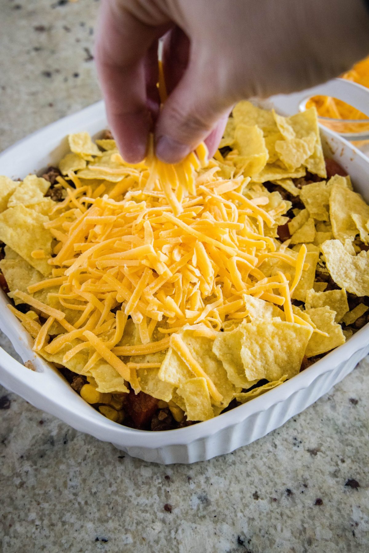 A hand sprinkling shredded cheddar cheese over the top of chips in a baking dish