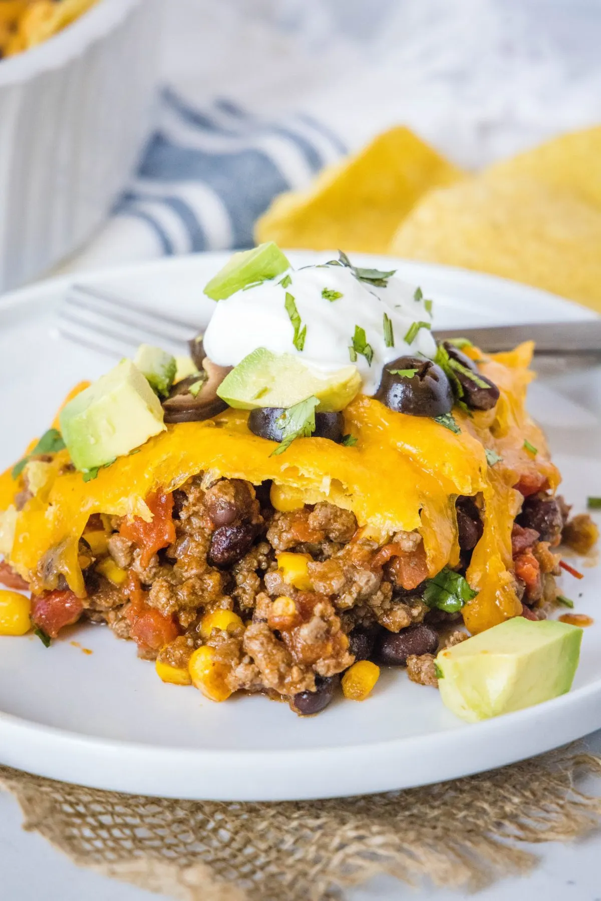 Ein Teller mit einer Portion Taco-Auflauf, garniert mit Sauerrahm, Koriander, Oliven und Avocado, im Hintergrund Chips