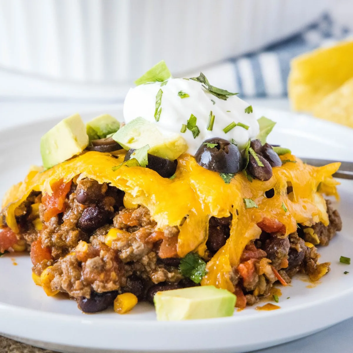 A serving of taco casserole on a plate, topped with sour cream, avocado, cilantro, and olives