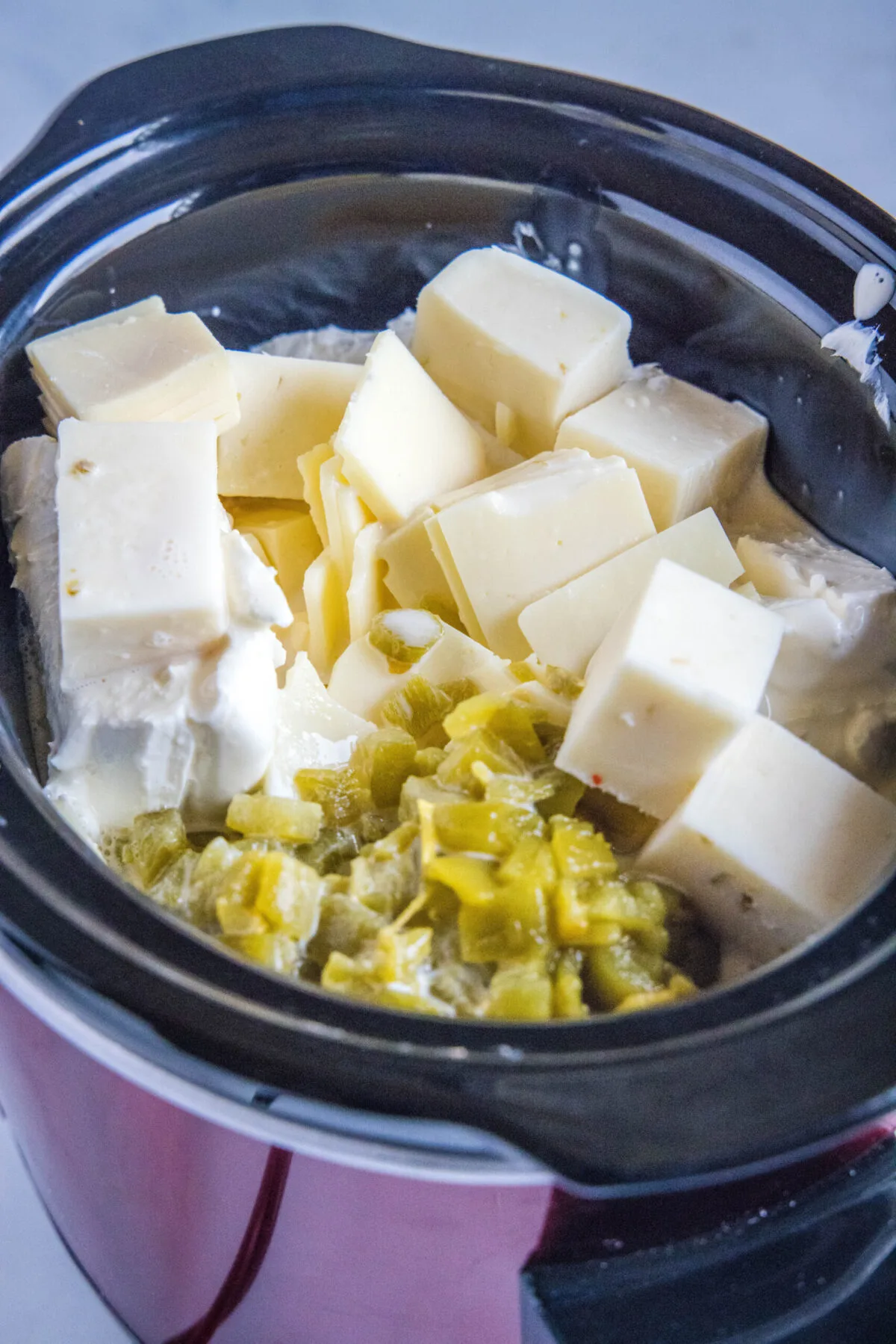 A crock pot filled with a block of cream cheese, cubes of American cheese and pepper jack cheese, and diced green chiles