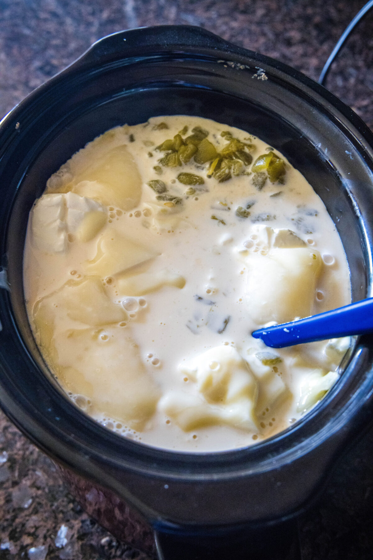 A rubber spatula stirring cheese, evaporated milk, and green chiles in a crock pot