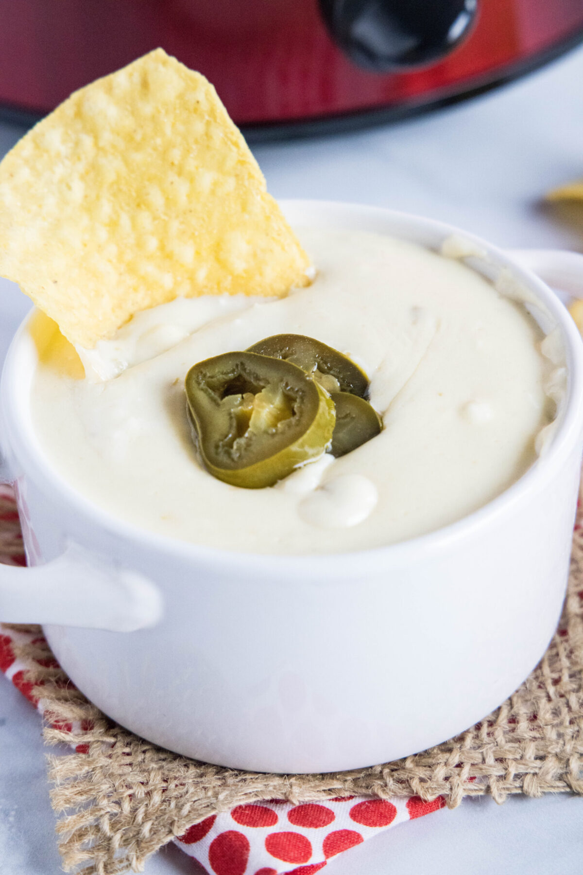 Overhead view of a bowl of white queso dip topped with pickled jalapeños with a chip in it, in front of a crock pot
