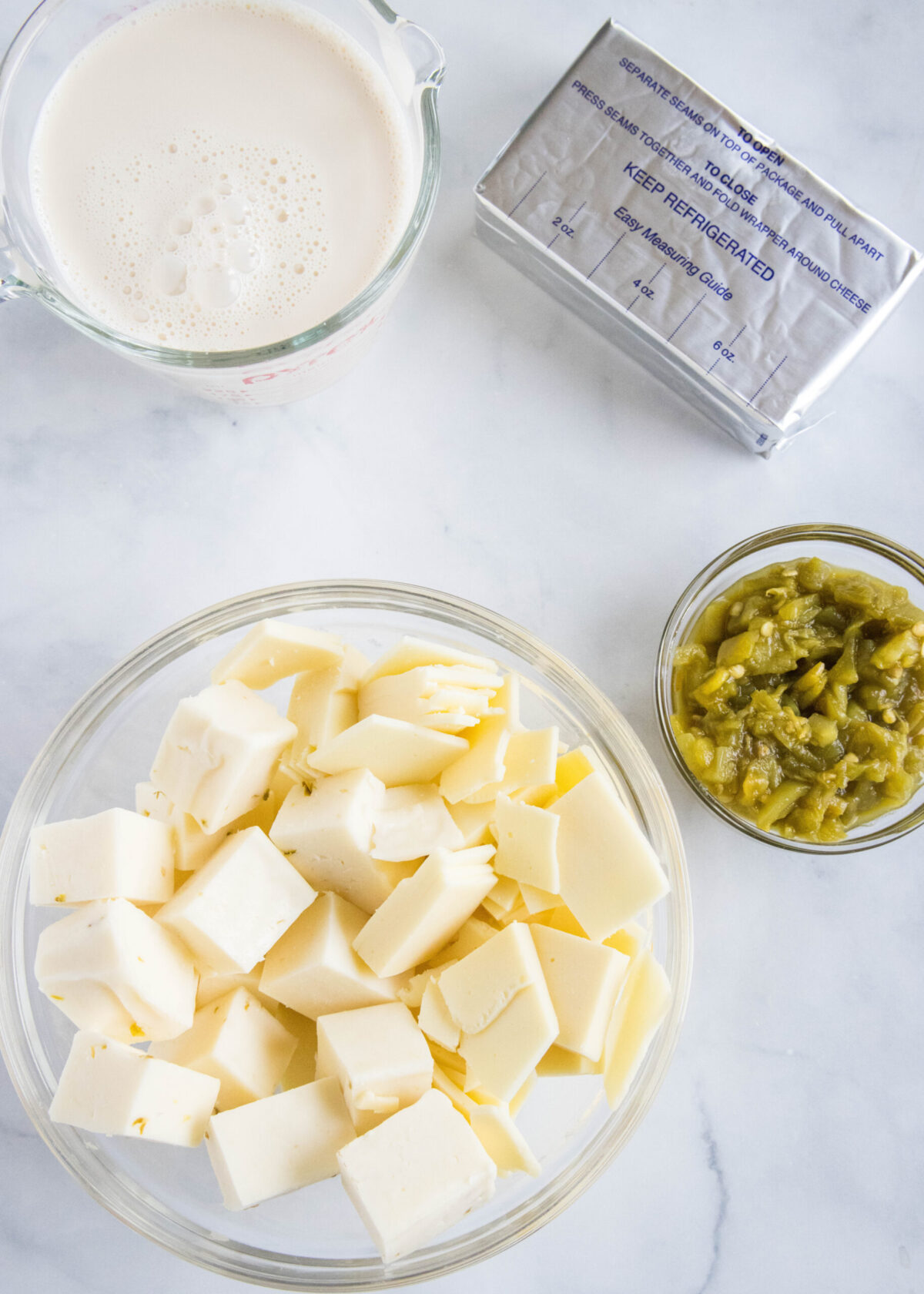 Overhead view of the ingredients needed for crock pot queso dip: a bowl of cubed American cheese and pepper jack cheese, a pyrex of evaporated milk, a pack of cream cheese, and a bowl of diced green chiles