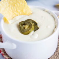 Close up of a bowl of white queso dip, topped with pickled jalapeños, with a chip sticking in it