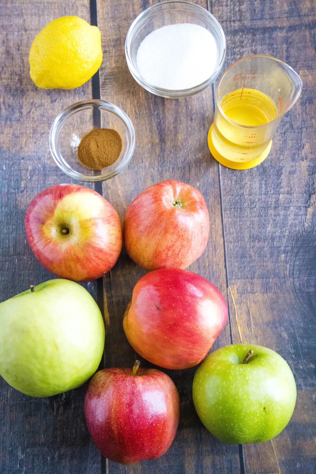 Overhead view of the ingredients needed for homemade applesauce: a variety of apples, a lemon, a bowl of cinnamon, a bowl of sugar, and a glass of water