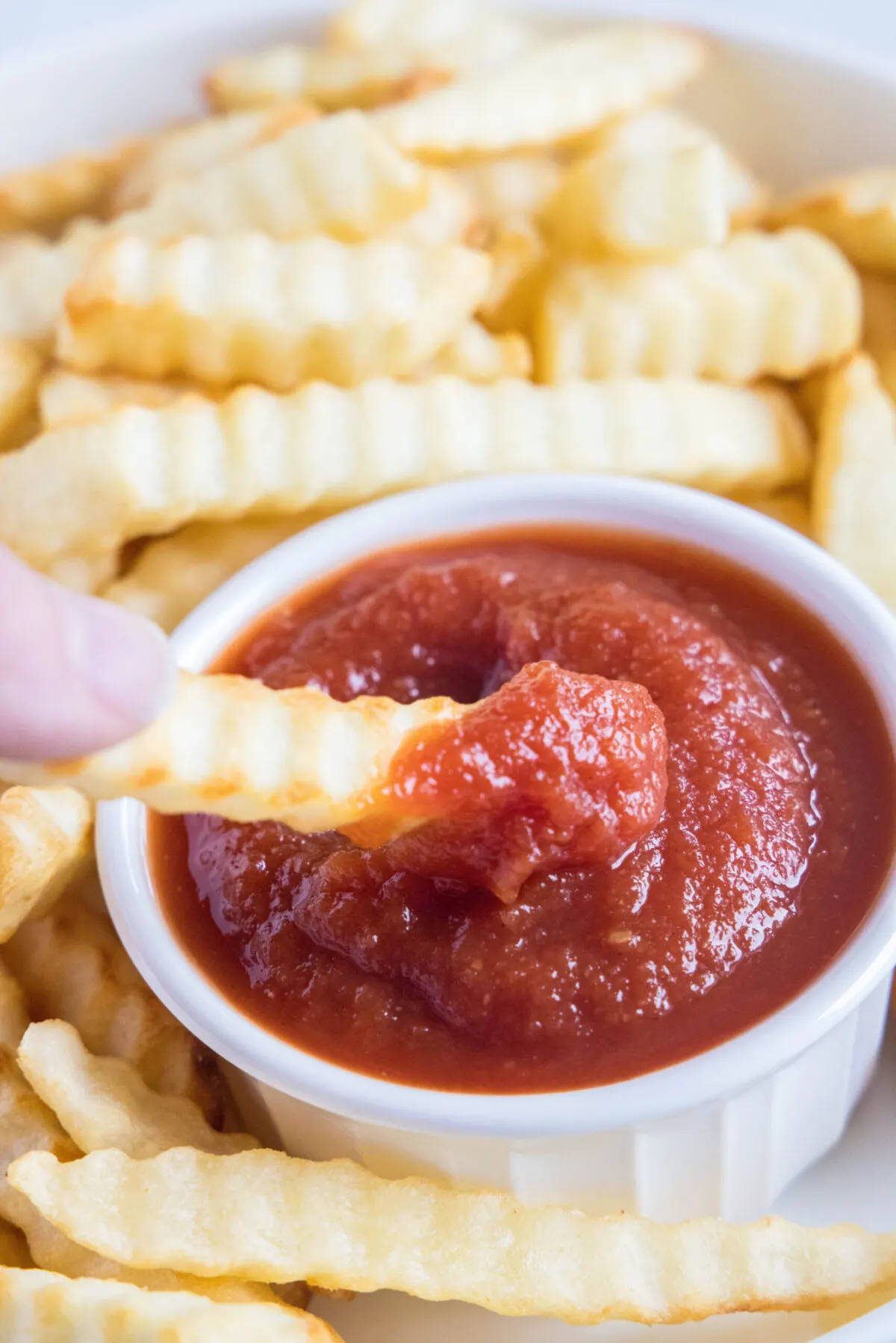 A hand dipping a fry into a ramekin of ketchup, surrounded by more fries