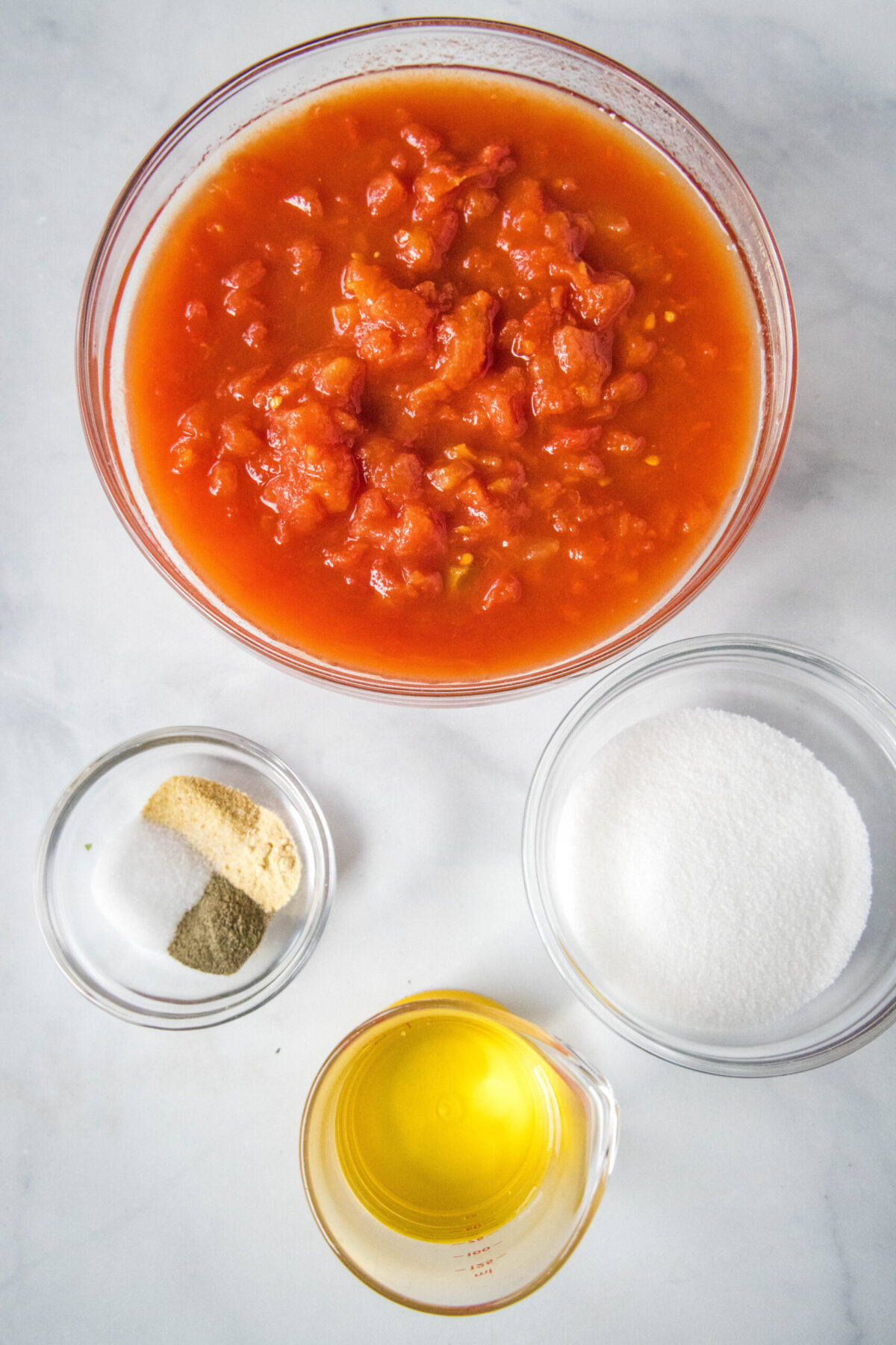 Overhead view of the ingredients needed for homemade ketchup: a bowl of canned tomatoes, a bowl of salt, pepper, onion powder, and garlic powder, a bowl of sugar, and a glass of vinegar