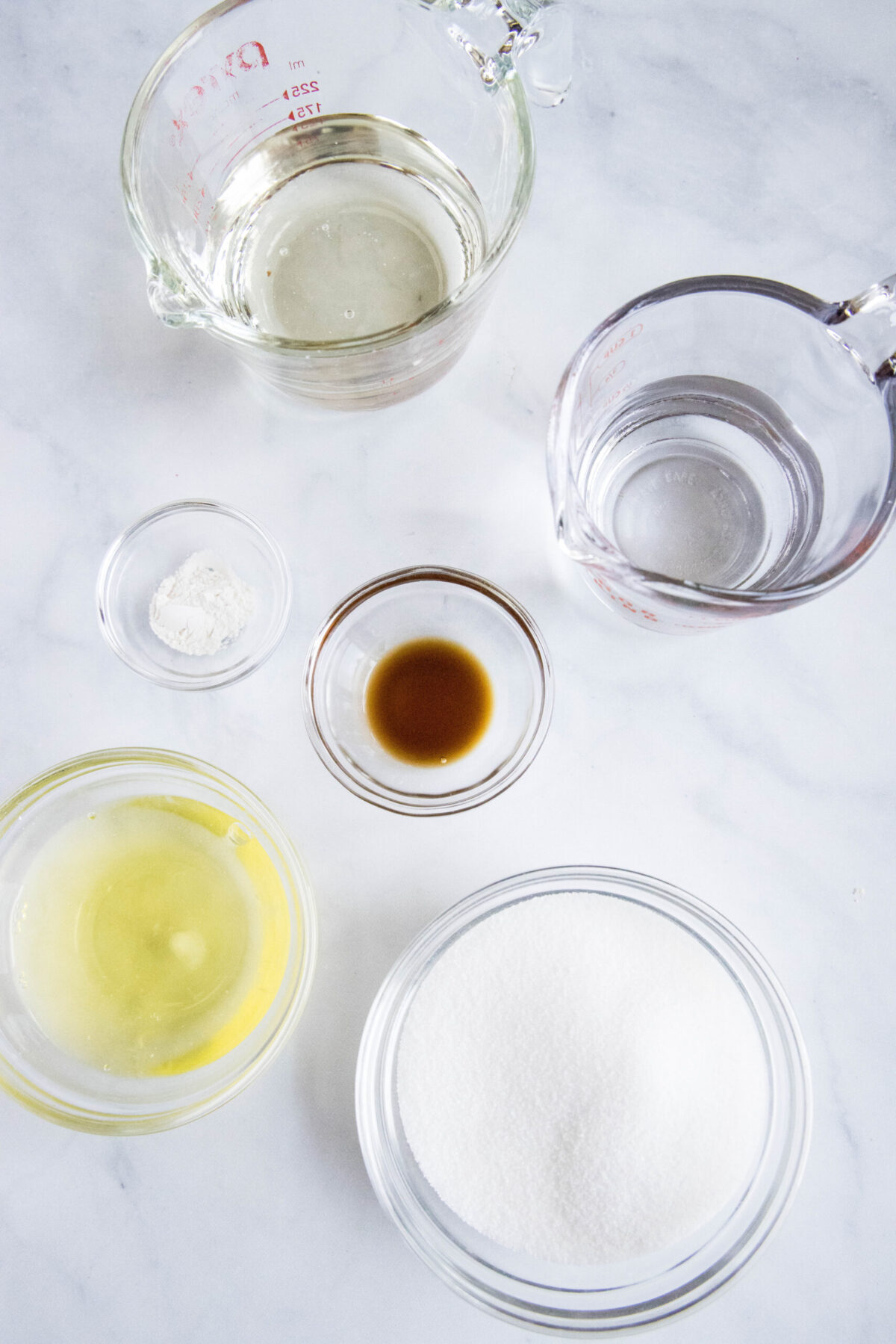 Overhead view of the ingredients needed for marshmallow fluff: a bowl of egg whites, a bowl of sugar, a pyrex of water, a bowl of vanilla, a pyrex of corn syrup, and a bowl of cream of tartar