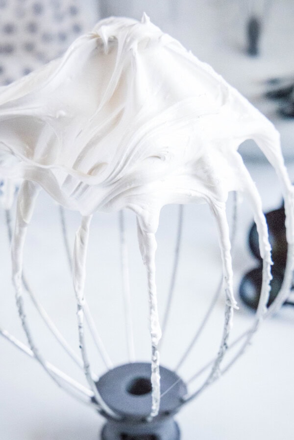 Close up of a stand mixer whisk attachment with stiff peaks of marshmallow fluff, with measuring spoons in the background