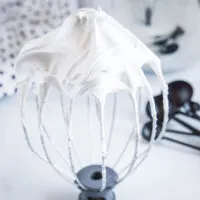 Close up of a stand mixer whisk attachment with stiff peaks of marshmallow fluff, with measuring spoons in the background