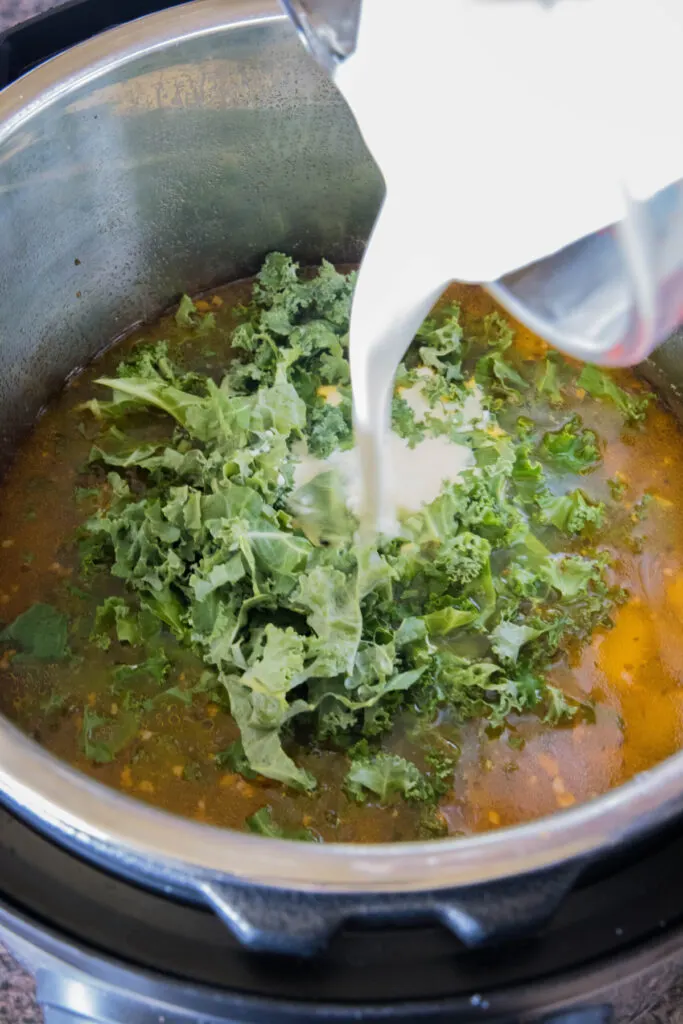 Heavy cream being poured on top of raw kale on top of a Zuppa Toscana in a pot