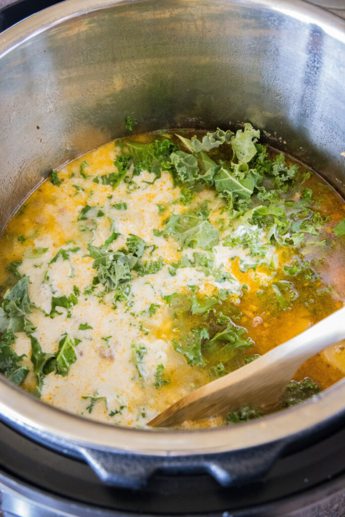 A wooden spoon stirring kale and cream into a pot of Zuppa Toscana