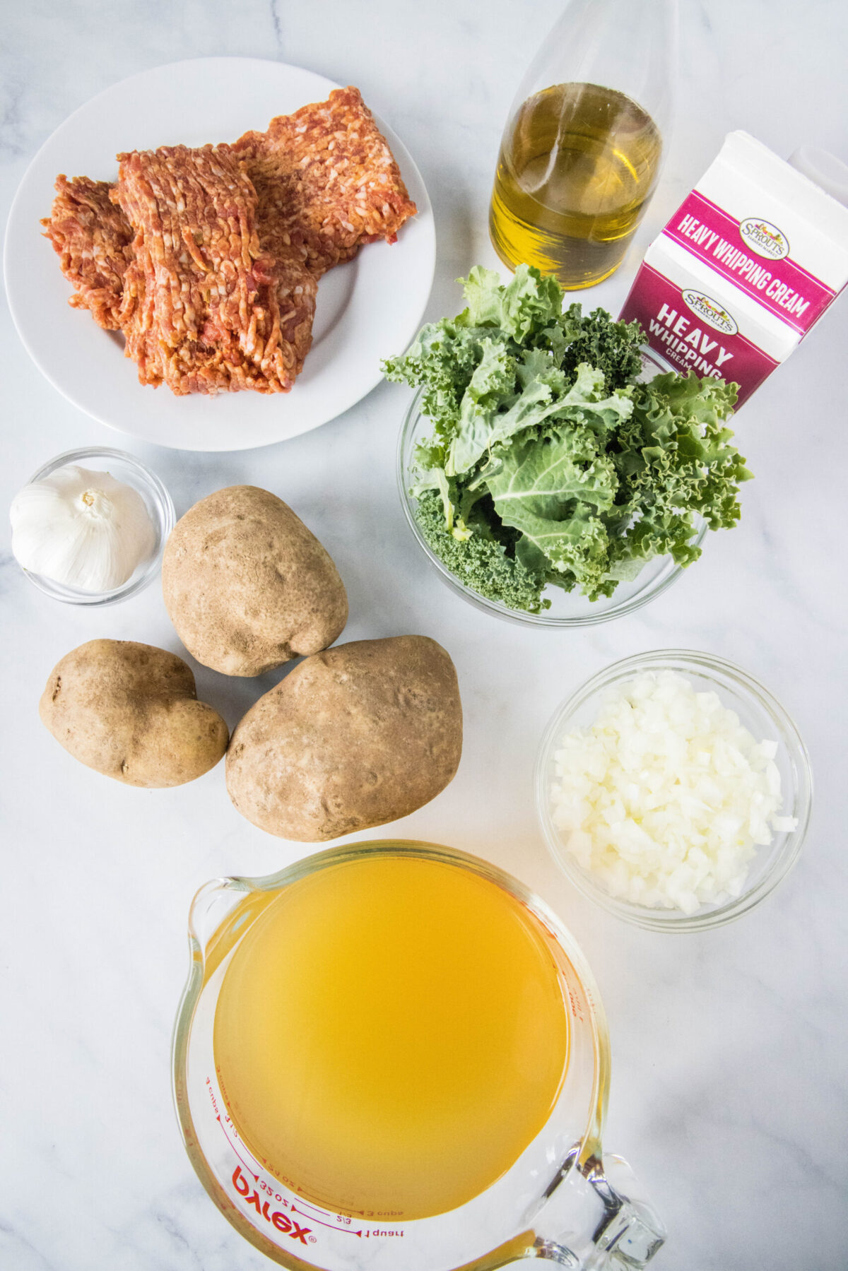 Overhead view of the ingredients needed for Zuppa Toscana: a plate of Italian sausage, a bowl of kale, a bowl of minced onion, a pyrex of chicken broth, a bowl of garlic, three potatoes, a jug of olive oil, and a carton of cream