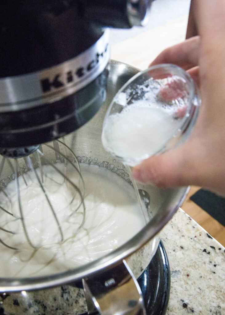 A hand pouring a bowl of gelatin and water into a stand mixer filled with cream