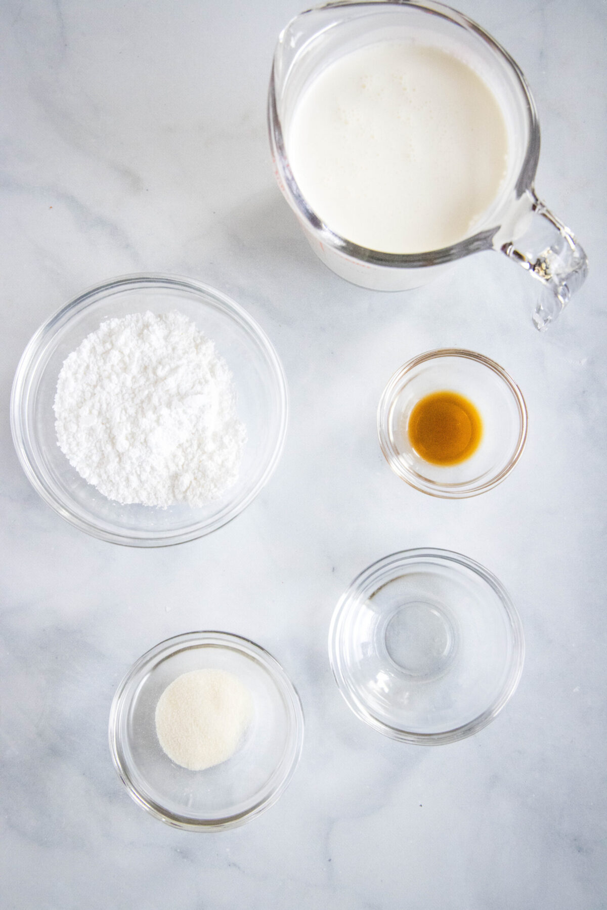 Overhead view of the ingredients needed for stabilized whipped cream: a pyrex of whipping cream, a bowl of powdered sugar, a bowl of vanilla extract, a bowl of gelatin powder, and a bowl of water
