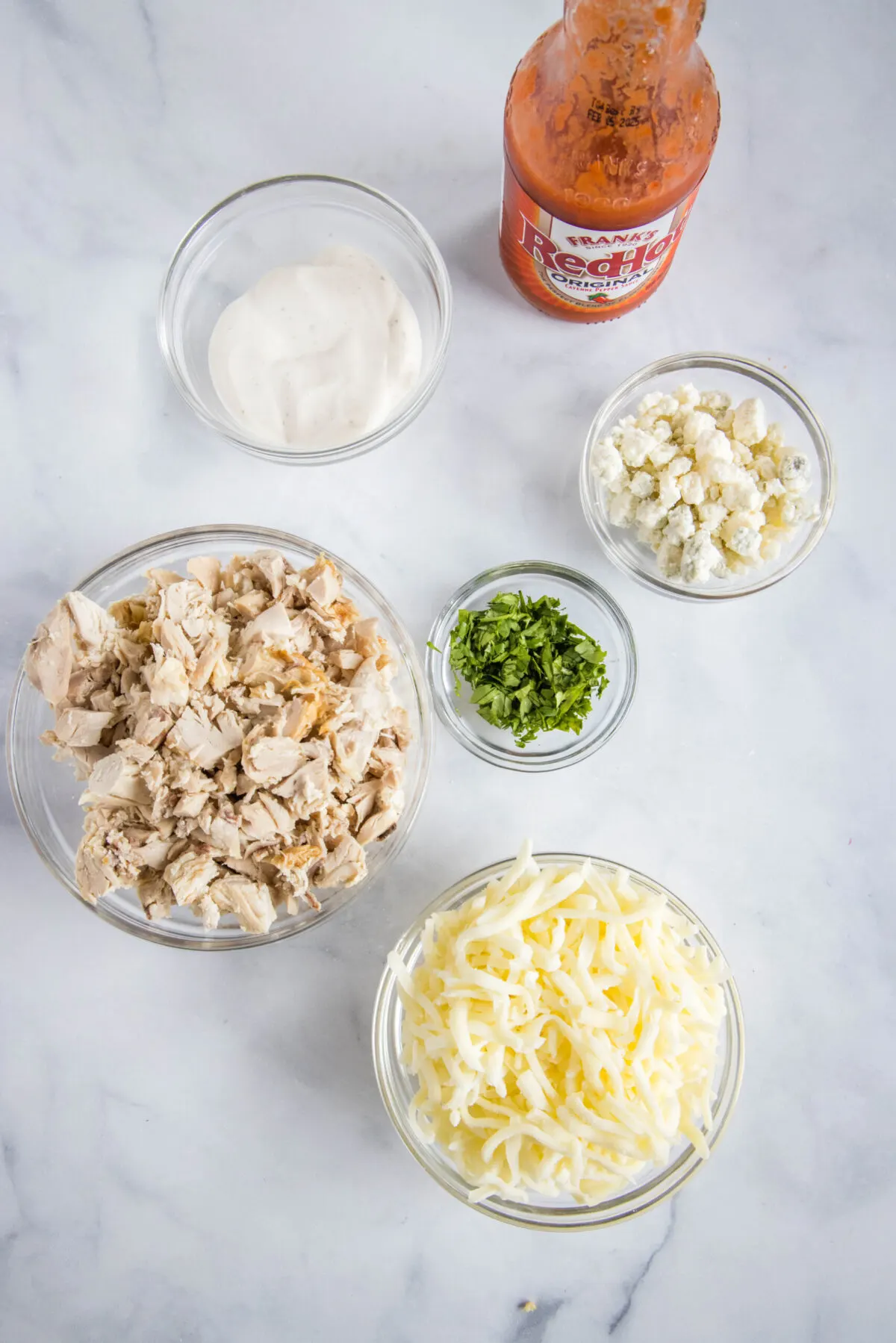 Overhead view of the ingredients needed for buffalo chicken pizza: a bowl of shredded chicken, a bowl of shredded cheese, a bowl of ranch dressing, a bowl of blue cheese, a bowl of parsley, and a bottle of wing sauce