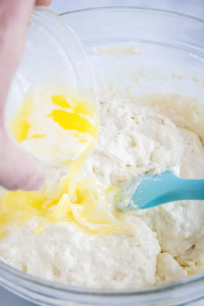 A hand pouring melted butter into a bowl with pancake batter and a spatula