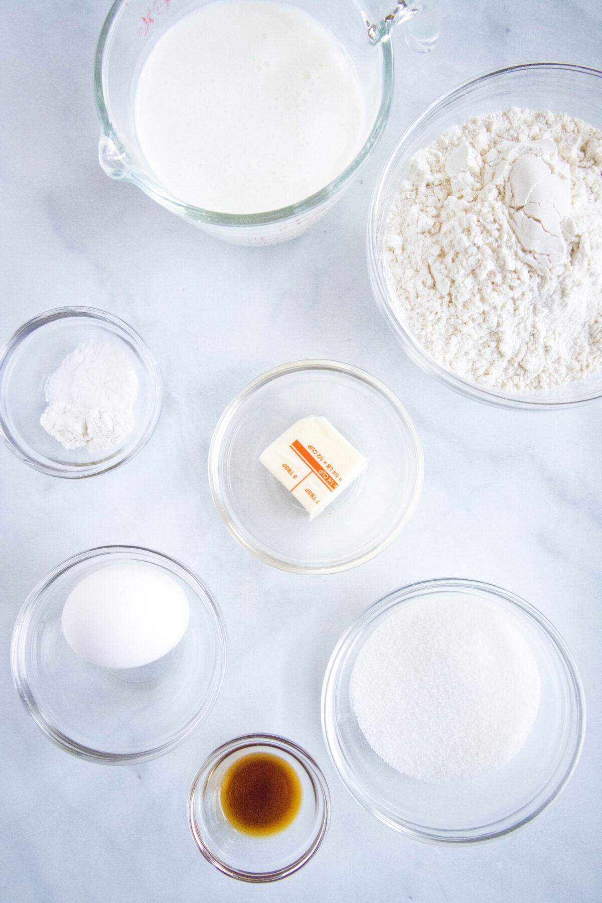 Overhead view of the ingredients needed for buttermilk pancakes: a bowl of flour, a pyrex of buttermilk, a bowl of sugar, a bowl of vanilla, a bowl of baking powder and soda, a cube of butter, and an egg.