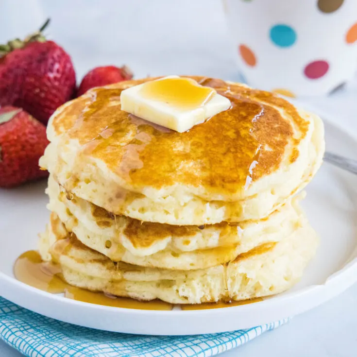 A stack of pancakes on a plate, topped with butter and syrup, with strawberries in the back.