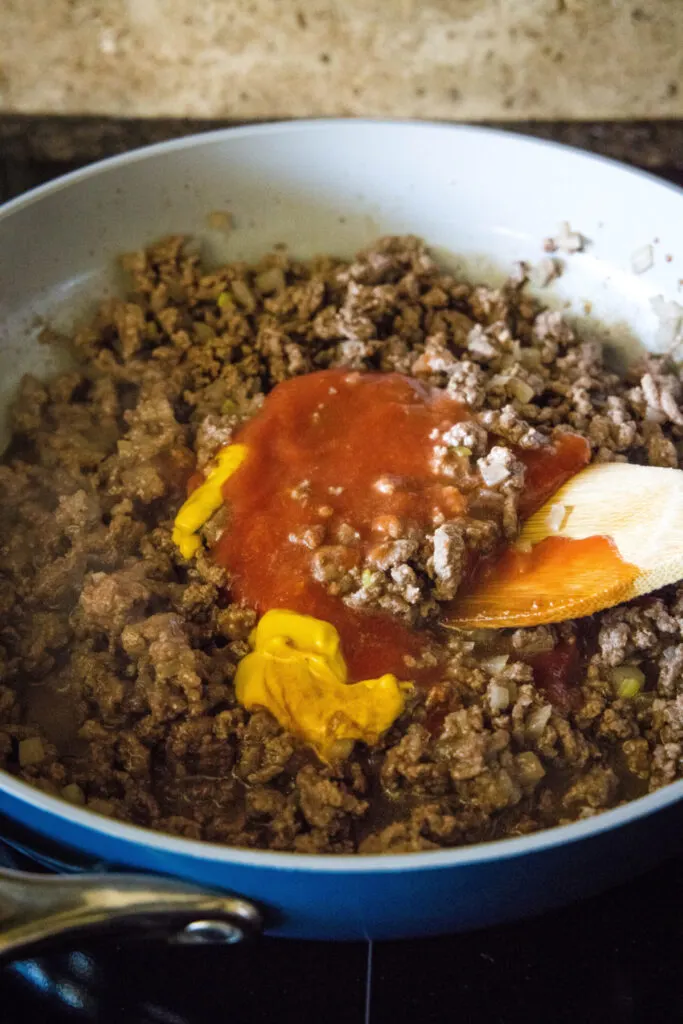 Mustard and Worcestershire sauce on top of cooked ground beef in a skillet, with a wooden spoon