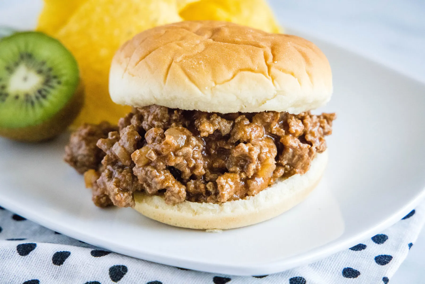 A sloppy joe sandwich on a plate with chips and a kiwi