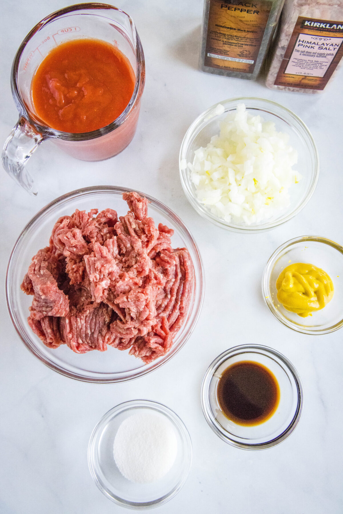 Overhead view of the ingredients needed for sloppy joes: a bowl of ground beef, a pyrex of tomato sauce, a bowl of onions, a bowl of mustard, a bowl of Worcestershire sauce, a bowl of sugar, a salt grinder, and a pepper grinder
