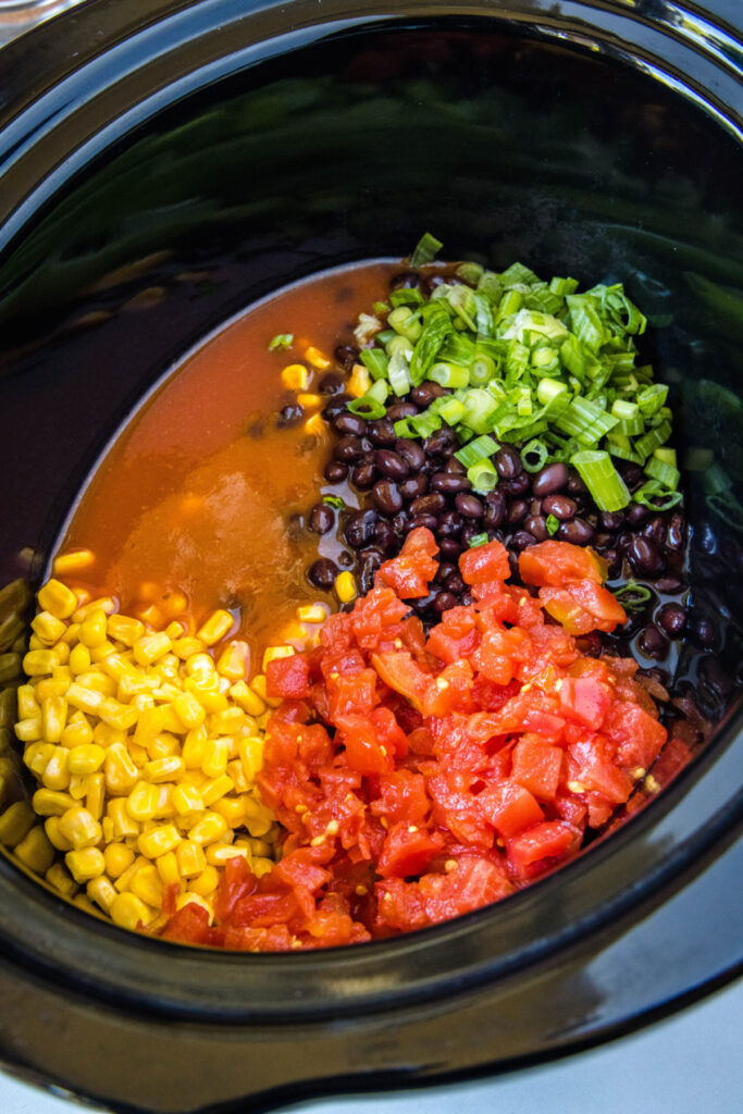 Overhead view of a crock pot full of diced tomatoes, corn, black beans, green onions, and enchilada sauce