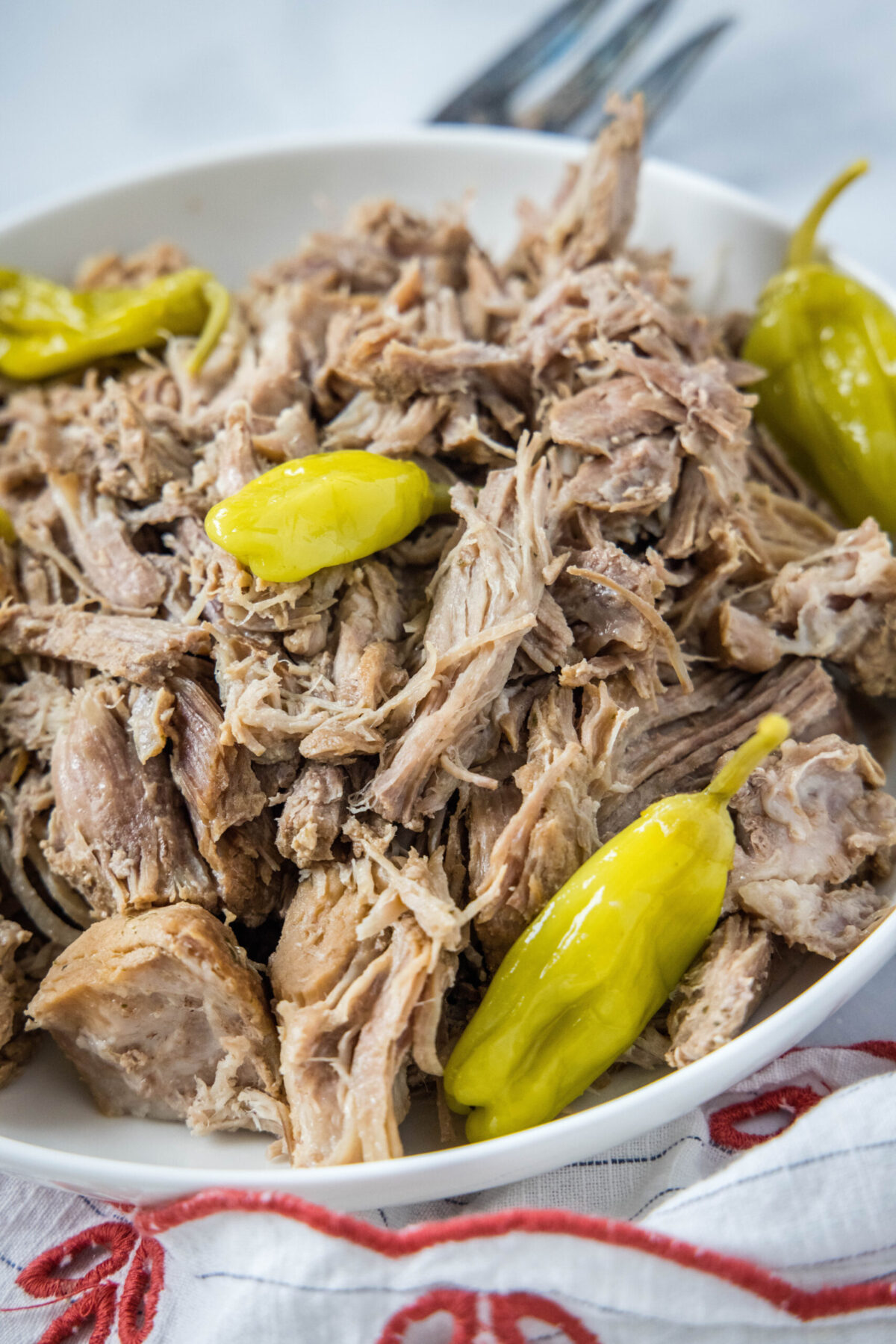 A bowl of shredded pork with pepperoncini peppers, next to a fork