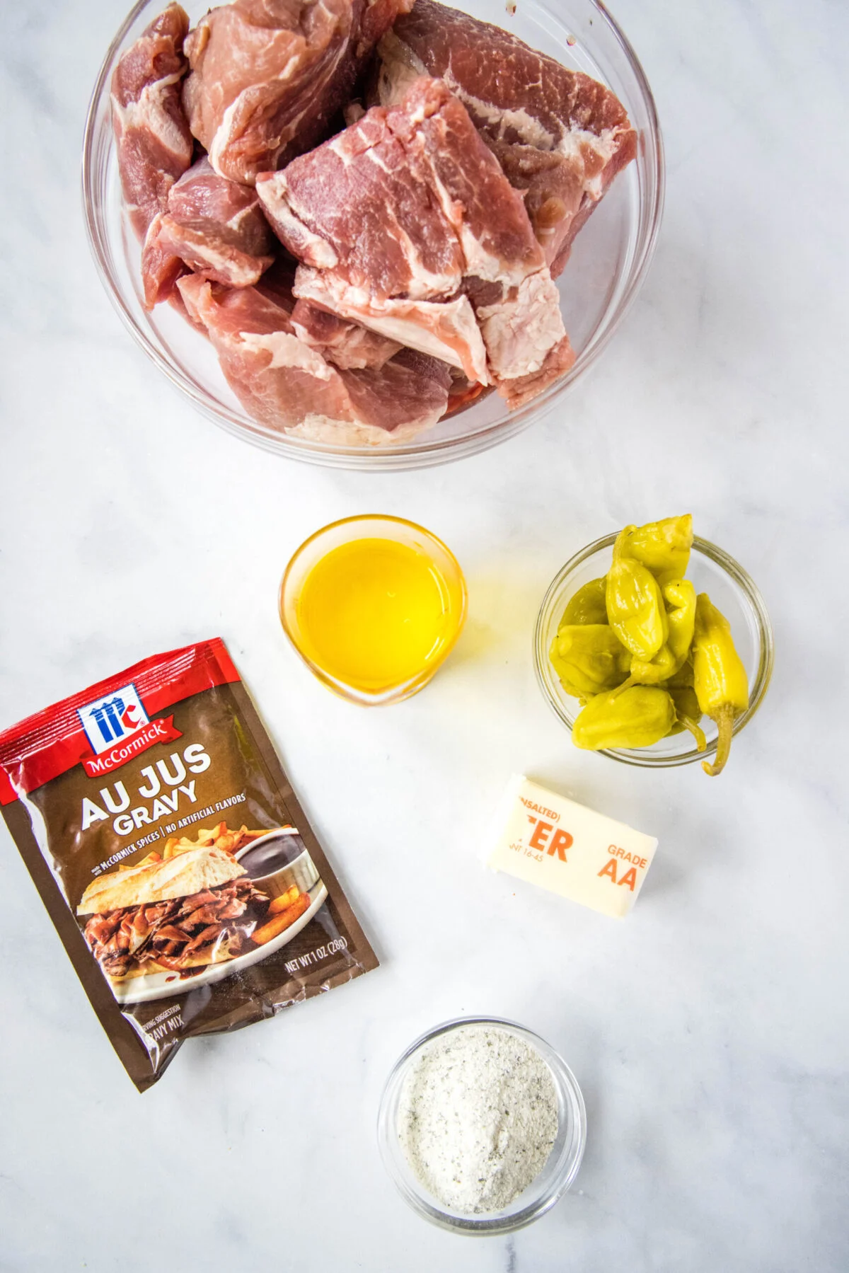 Overhead view of the ingredients needed for Mississippi pork roast: a bowl of pork shoulder, a bowl of pepperoncinis, a bowl of pepperoncini juice, a bowl of ranch dressing mix, a packet of au jus, and a stick of butter