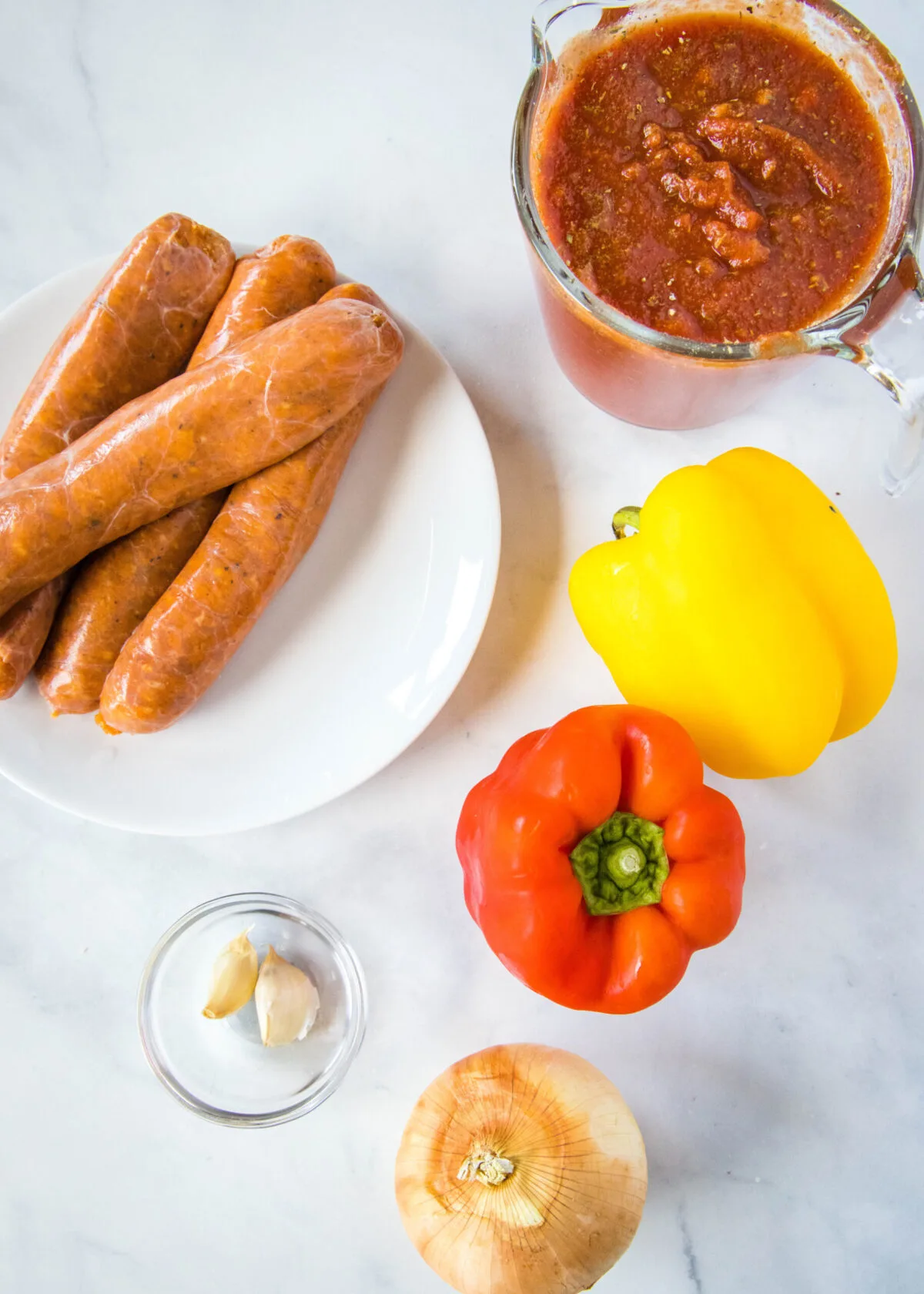 Overhead view of the ingredients needed for crock pot sausage and peppers: a plate of chicken sausage, a jar of marinara sauce, a yellow bell pepper, a red bell pepper, a yellow onion, and some garlic cloves