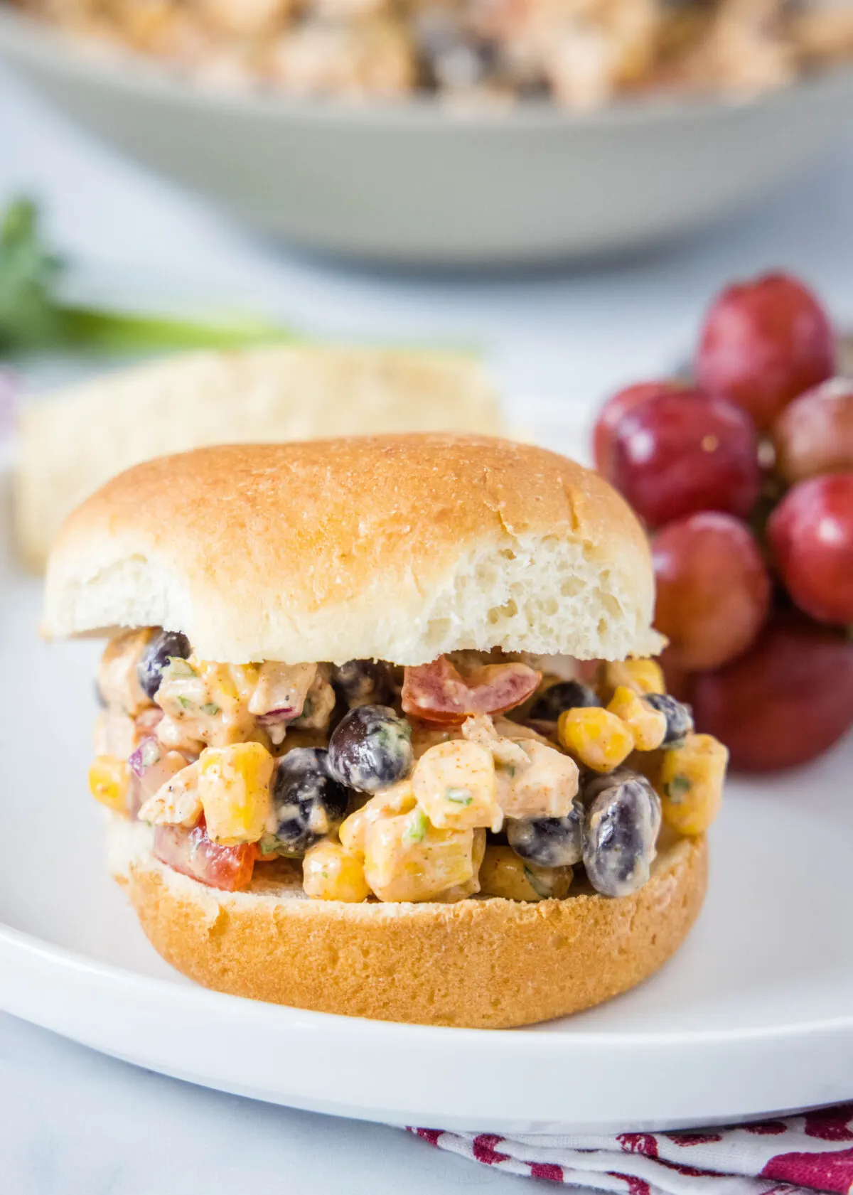 A burger bun filled with chicken salad with corn and beans, on a plate next to some grapes