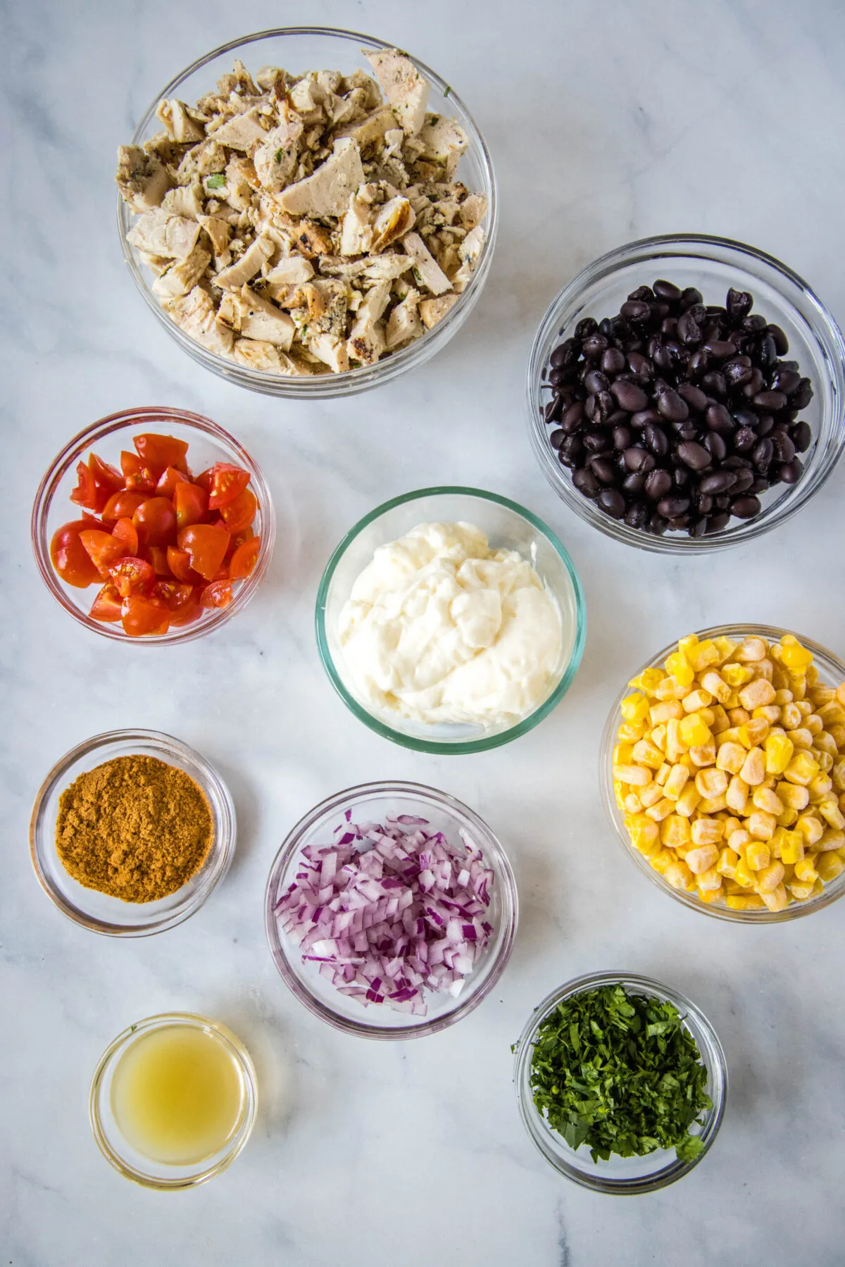 Overhead view of the ingredients needed for southwest chicken salad: a bowl of shredded chicken, a bowl of black beans, a bowl of chopped tomatoes, a bowl of mayo, a bowl of corn, a bowl of chopped onion, a bowl of cilantro, a bowl of taco seasoning, and a bowl of lime juice