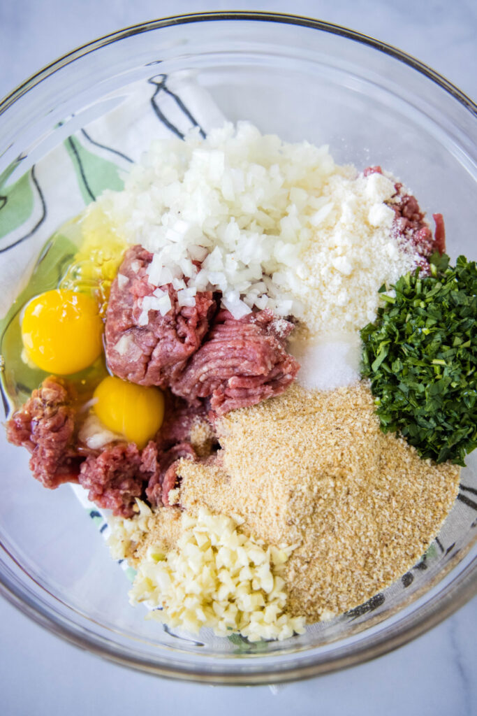 Overhead view of a mixing bowl with ground beef, onions, garlic, salt, breadcrumbs, parsley, eggs, milk, and olive oil.