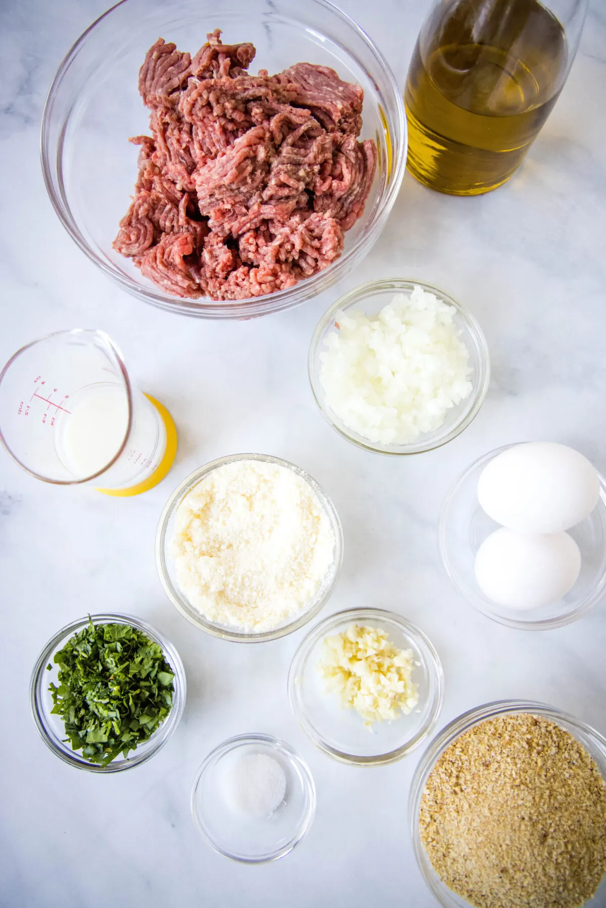 Overhead view of the ingredients needed for air fryer meatballs: a bowl of ground beef, a bowl of onions, a bowl of garlic, a bowl of parsley, a bowl of breadcrumbs, a glass of milk, a bottle of olive oil, a bowl of parmesan cheese, a bowl of salt, and some eggs.