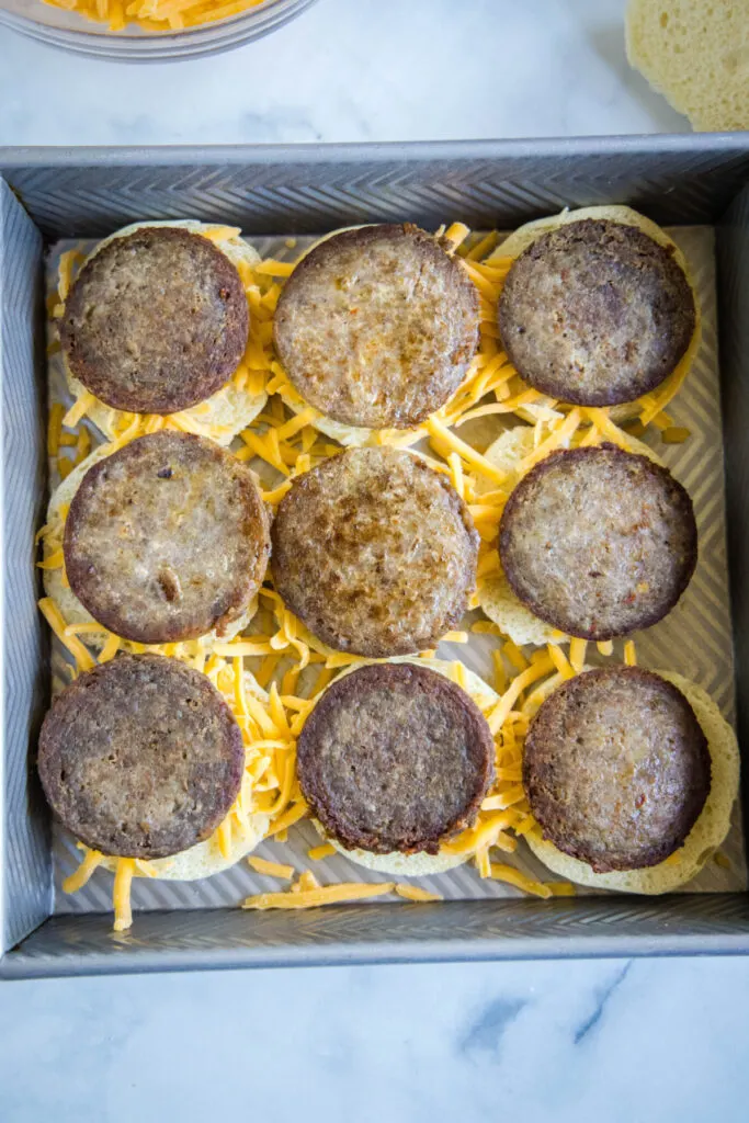Overhead view of a baking tray with nine sausage patties on top of cheese on top of slider buns