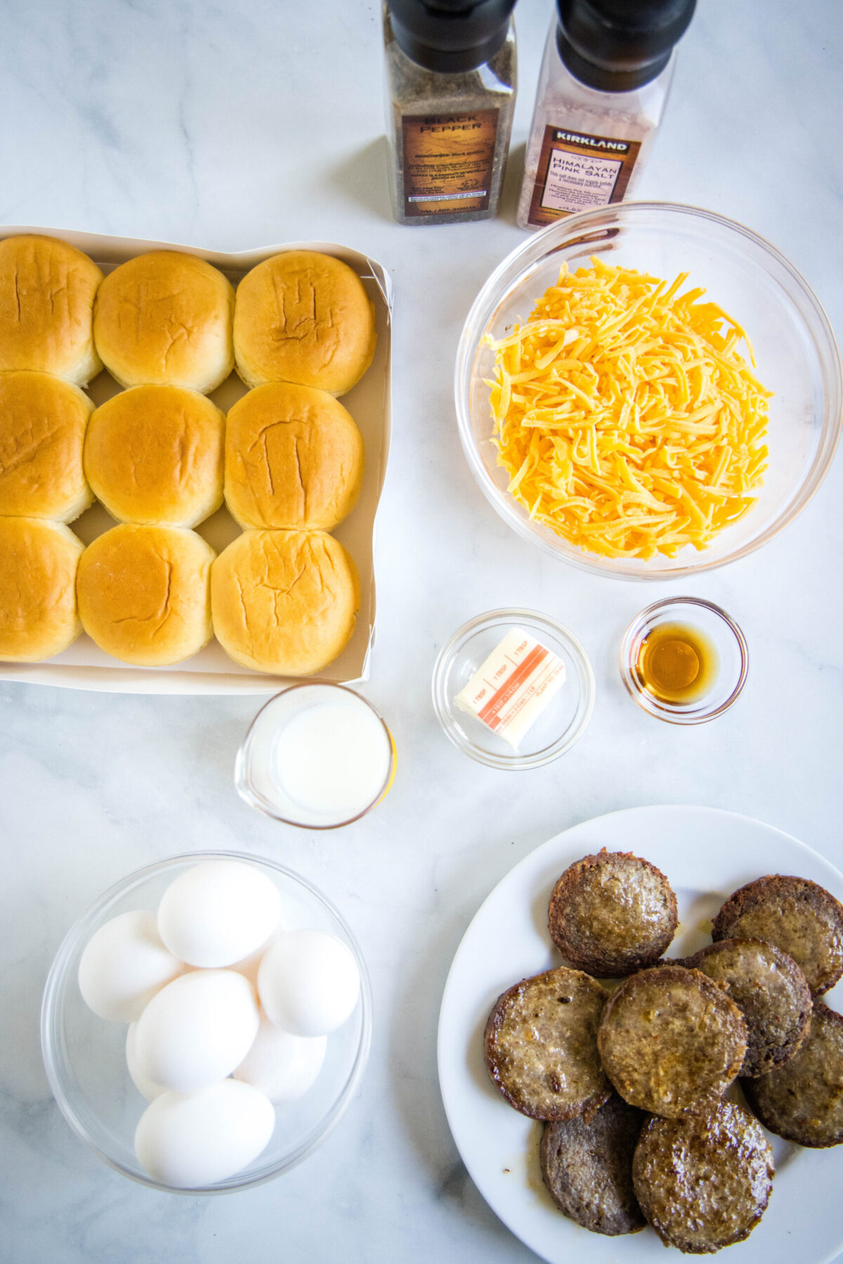 Overhead view of the ingredients needed for breakfast sliders: a pack of slider buns, a plate of sausages, a bowl of eggs, a bowl of shredded cheese, a bowl of milk, a bowl of maple syrup, some butter, and salt and pepper grinders